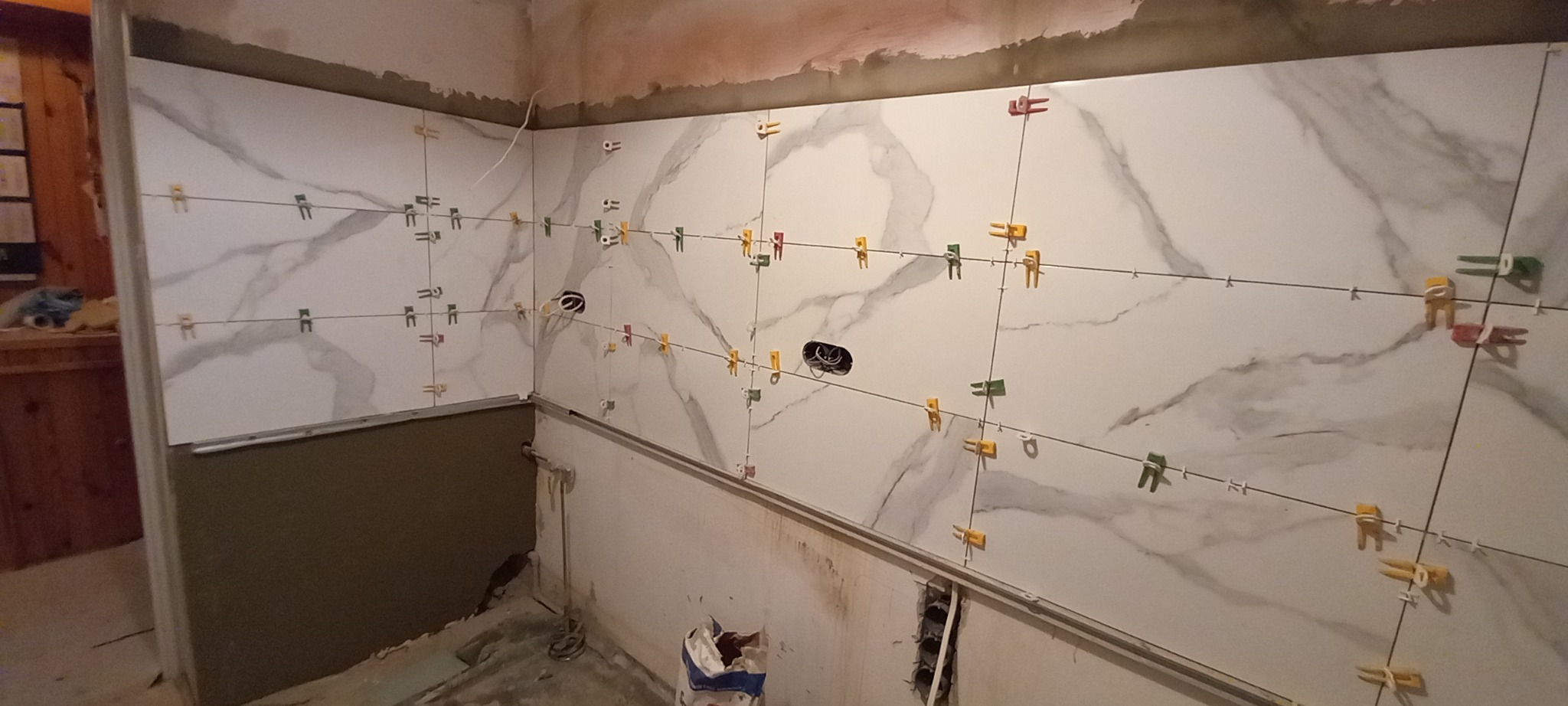 Ściana w trakcie układania płytek imitujących marmur, z widocznymi krzyżykami dystansowymi w różnych kolorach i przewodami elektrycznymi wystającymi ze ściany.