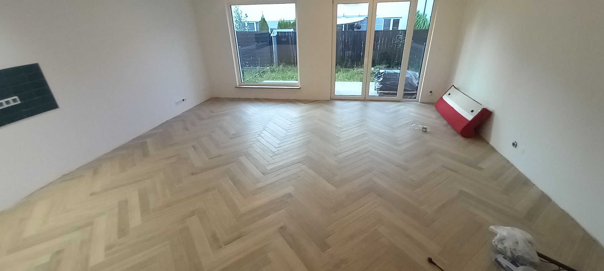 Jasny parkiet ułożony we wzór jodełki francuskiej w pustym pokoju z białymi ścianami i dużymi oknami balkonowymi, widoczne przygotowane materiały do wykończenia.