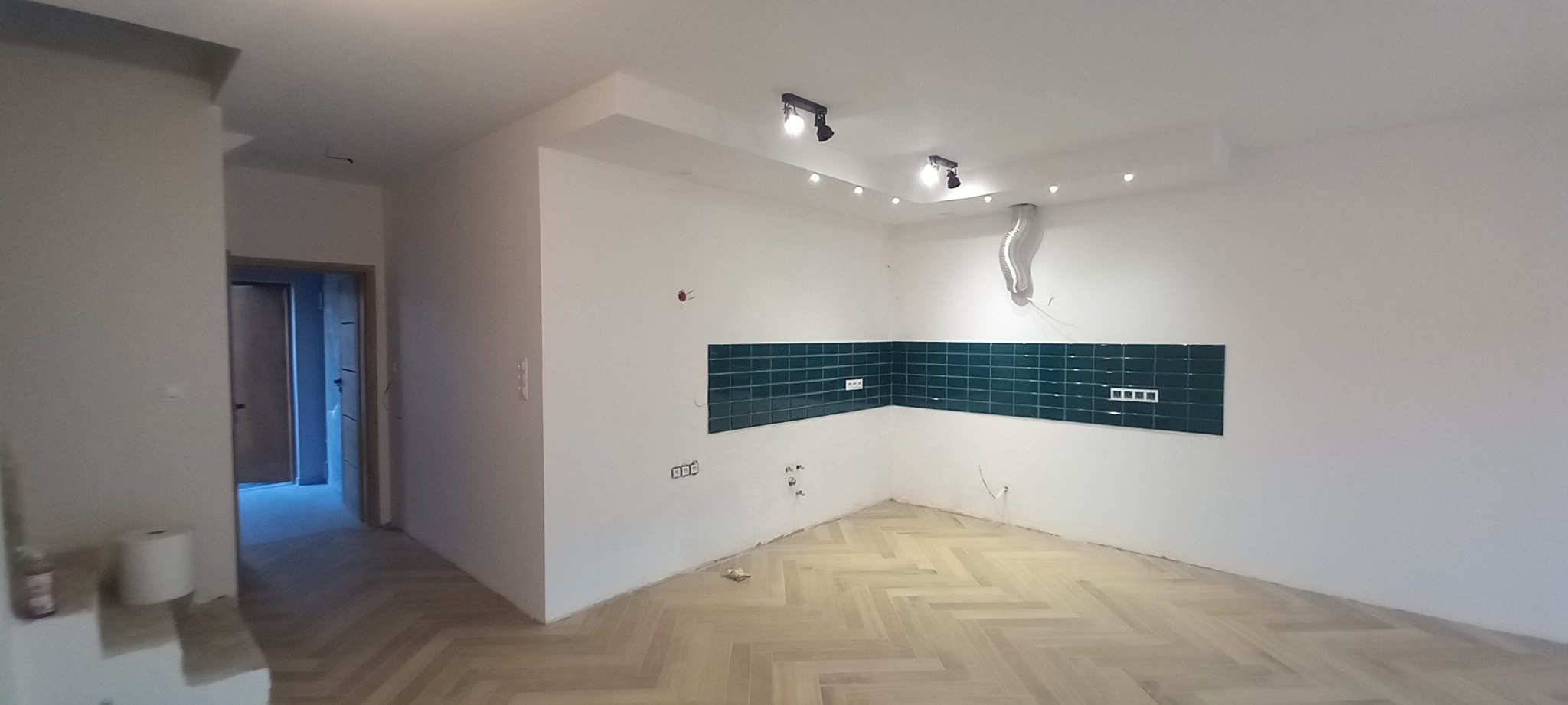 Wnętrze w trakcie remontu: ułożona podłoga w jodełkę, fragment ściany z zielonymi płytkami i gniazdkami elektrycznymi, widoczne przewody i rury wentylacyjne pod sufitem.