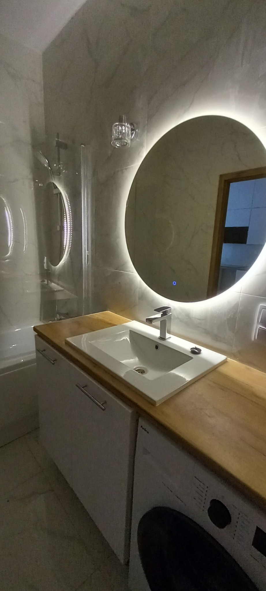 Nowoczesna łazienka z okrągłym lustrem podświetlanym LED, białą umywalką wpuszczoną w drewniany blat i pralką Amica w zabudowie.