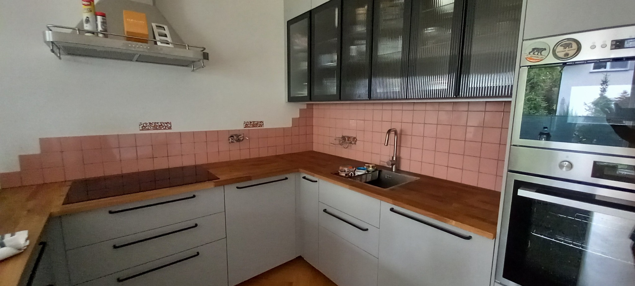 Fragment kuchni z różowymi płytkami ściennymi ułożonymi schodkowo, jasnoszare szafki z drewnianym blatem, czarne uchwyty, szklane szafki wiszące z czarnymi ramami, okap, piekarnik z naklejkami...