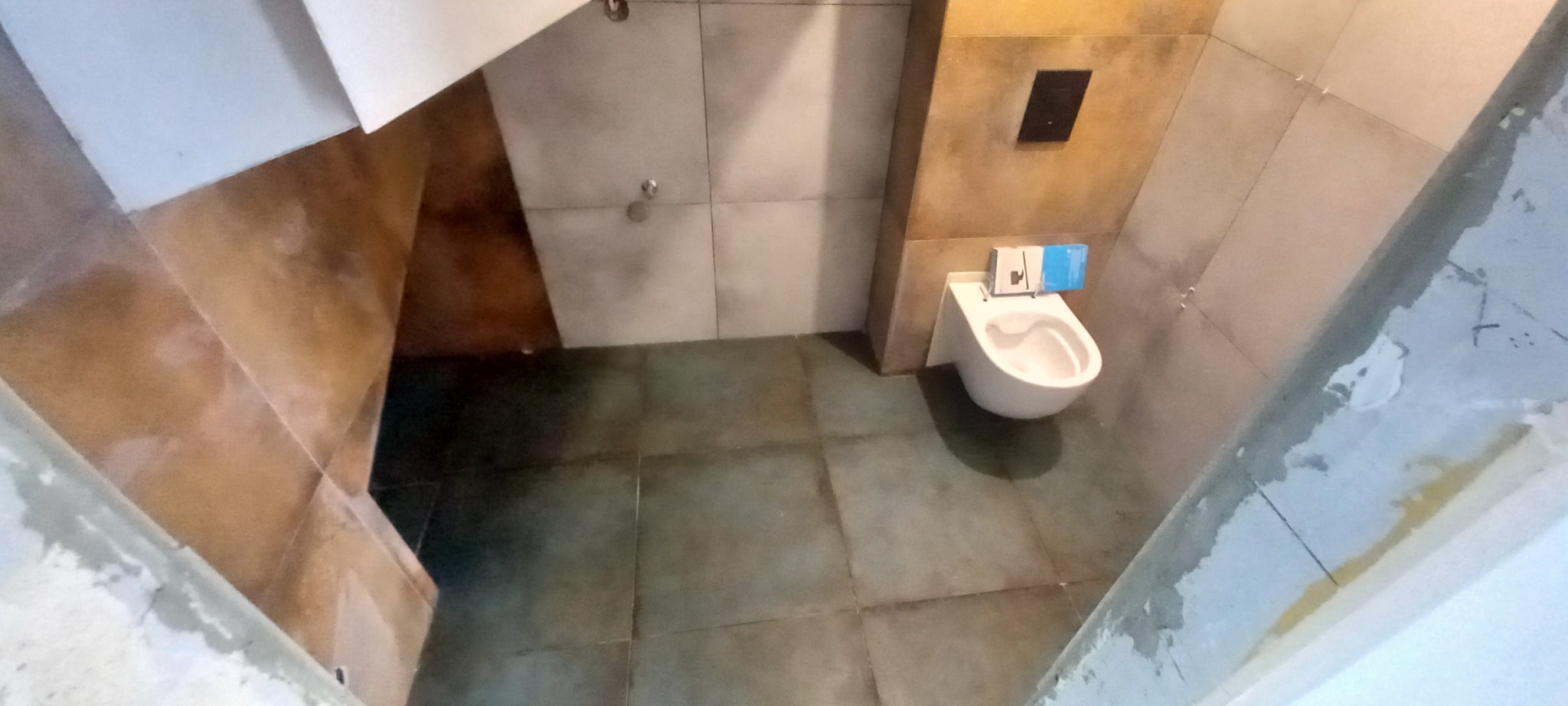 Niedokończona łazienka z zamontowaną toaletą wiszącą, widoczne surowe ściany i podłoga wymagające wykończenia, opakowania po produkcie na sedesie.