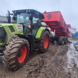 PDUH WOD-KAN-BUD Rafał Kołtuński - Zielony traktor Claas Arion z czerwoną przyczepą wywrotką stoi na błotnistym placu budowy, w tle widoczne materiały budowlane Ytong i Silka oraz budynek w trakcie wznoszenia.