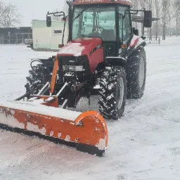 Czerwony traktor z pomarańczowym pługiem odśnieżnym na zasypanym śniegiem placu, w tle budynek i drzewa.