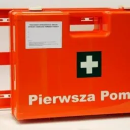 Pomarańczowa, plastikowa apteczka pierwszej pomocy K10 z białym napisem i zielonym krzyżem, obok widoczna pusta obudowa apteczki z uchwytami do montażu.