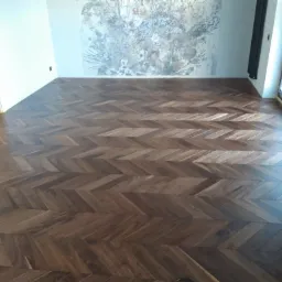 Ułożona podłoga z ciemnego drewna w jodełkę francuską, widok na ścianę z delikatnym, rozmytym wzorem kwiatowym i grzejnikiem w tle.