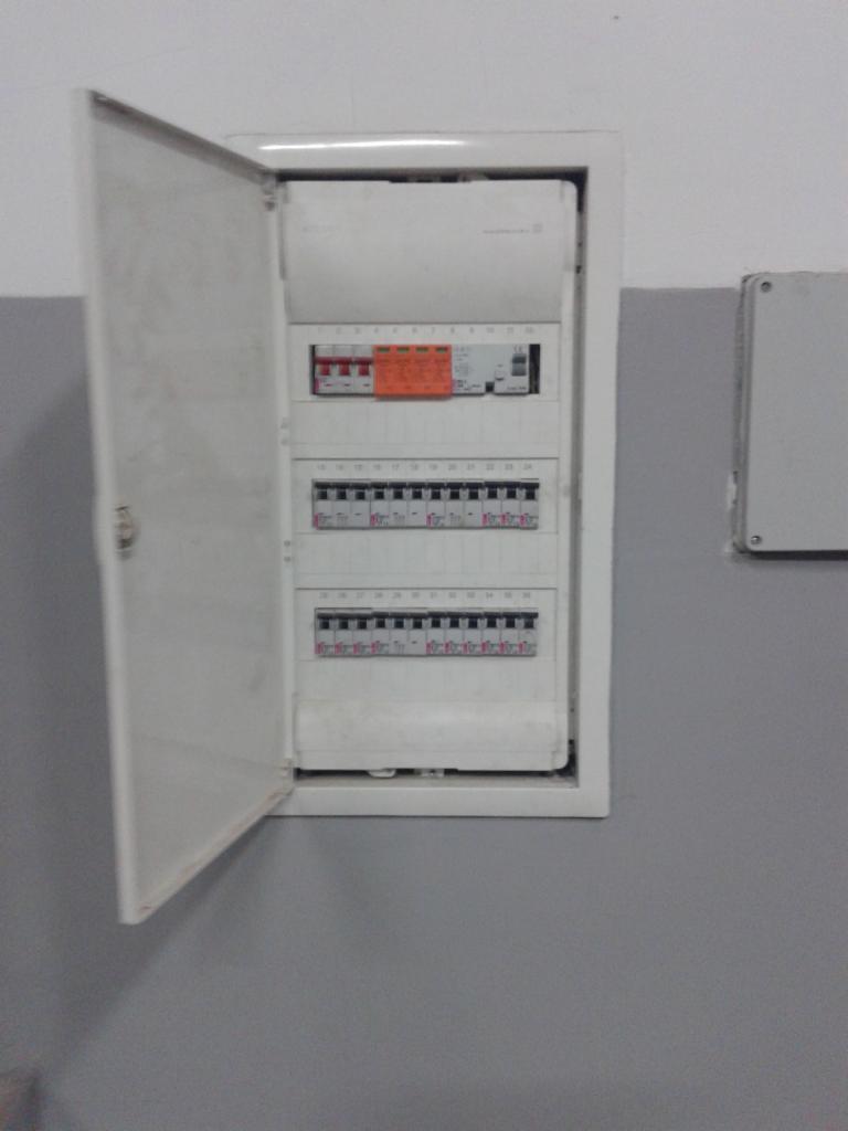 Otwarta skrzynka elektryczna z bezpiecznikami na szarej ścianie, widoczne trzy rzędy zabezpieczeń i naklejka ostrzegawcza. Obok puszka instalacyjna.