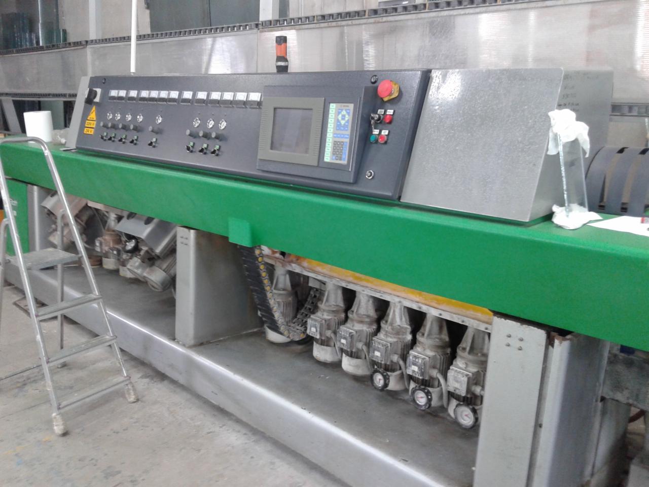 Szara maszyna przemysłowa z zielonym blatem, wyposażona w panel sterowania z ekranem i przyciskami. Widoczne silniki i łańcuchy. Obok drabina.