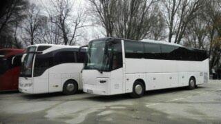 Dwa białe autokary turystyczne zaparkowane na placu, widoczne drzewa w tle, jeden z autokarów z czarnym dachem.