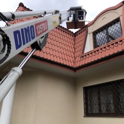 RAMZES Technologie Diamentowe Paweł Purzycki - Praca na wysokościach przy użyciu podnośnika koszowego DINO 135TB II, widoczny fragment dachu z brązową dachówką i okno z kratą w domu o beżowej elewacji.