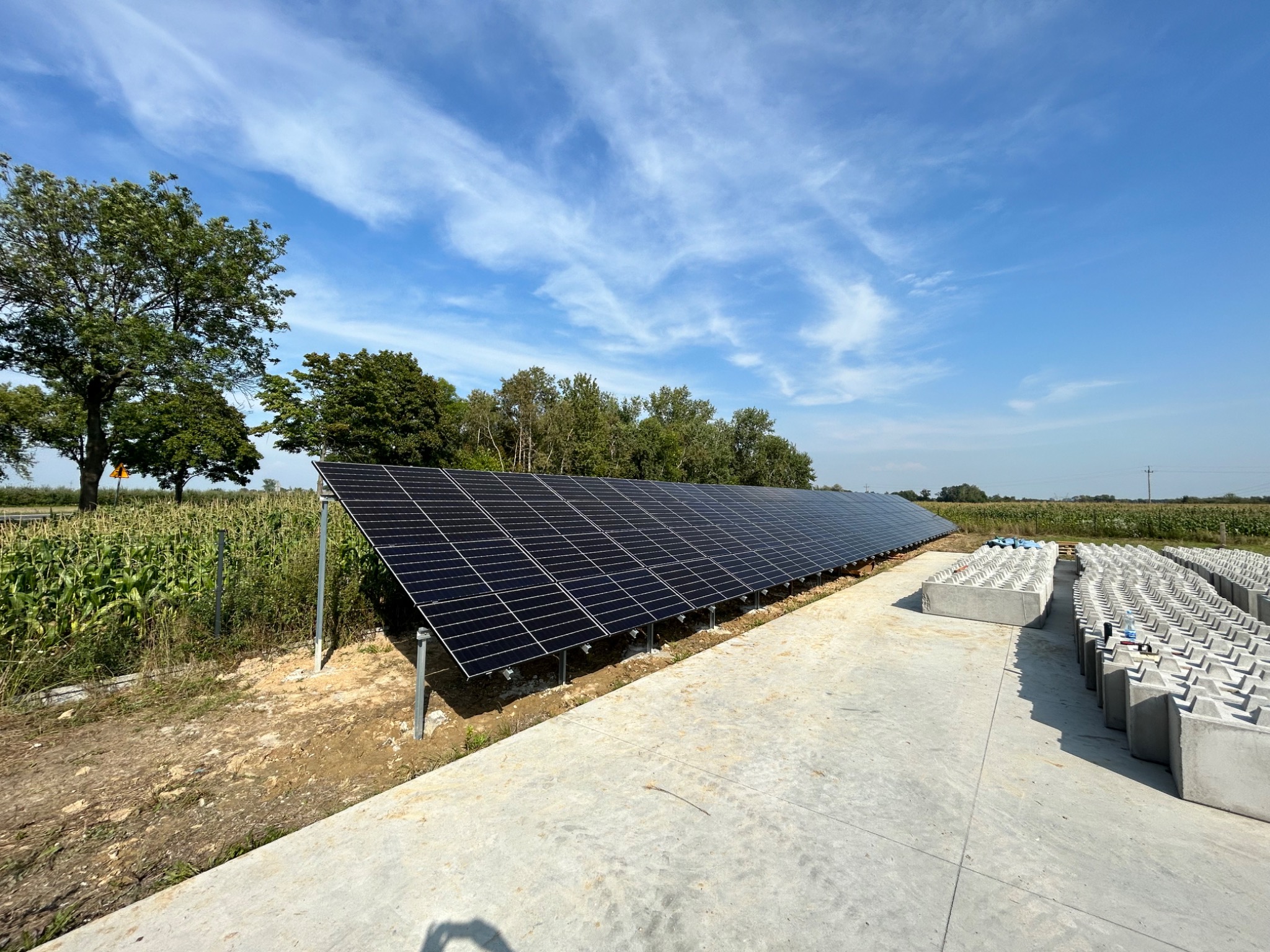 Długa instalacja paneli słonecznych w rzędzie na betonowej powierzchni, z widocznymi betonowymi blokami balastowymi, w tle pole uprawne i drzewa pod błękitnym niebem.