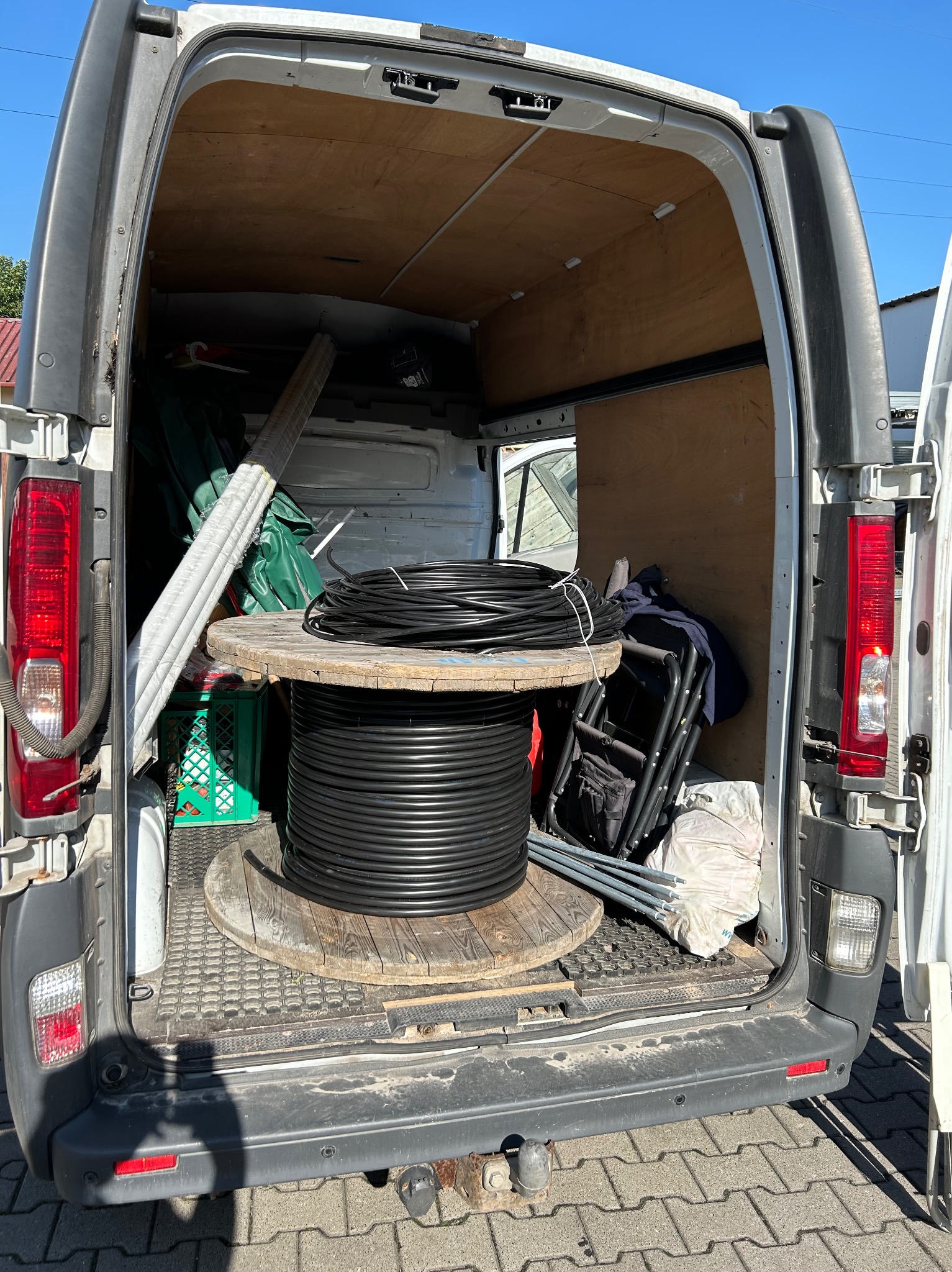 Bęben z czarnym kablem elektrycznym w bagażniku busa firmy instalacyjnej w Warszawie, obok drabina i inne akcesoria.