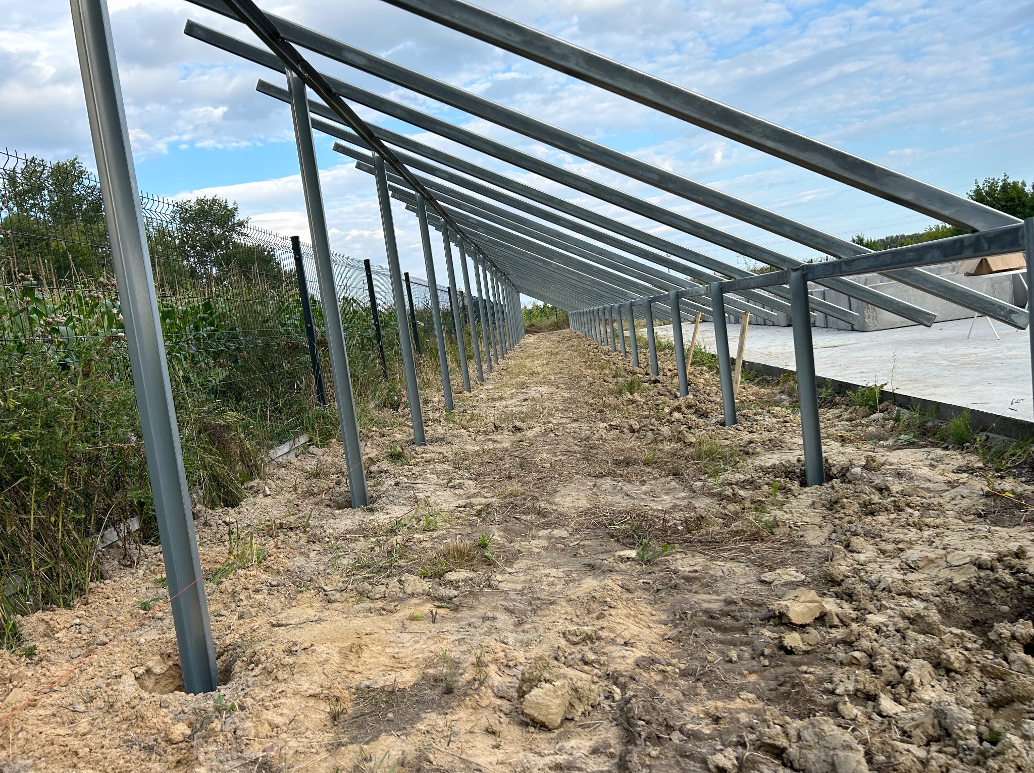 Metalowa konstrukcja wsporcza dla paneli słonecznych w trakcie montażu na gruncie, widoczne słupki i poprzeczki, w tle zieleń i błękitne niebo z chmurami.