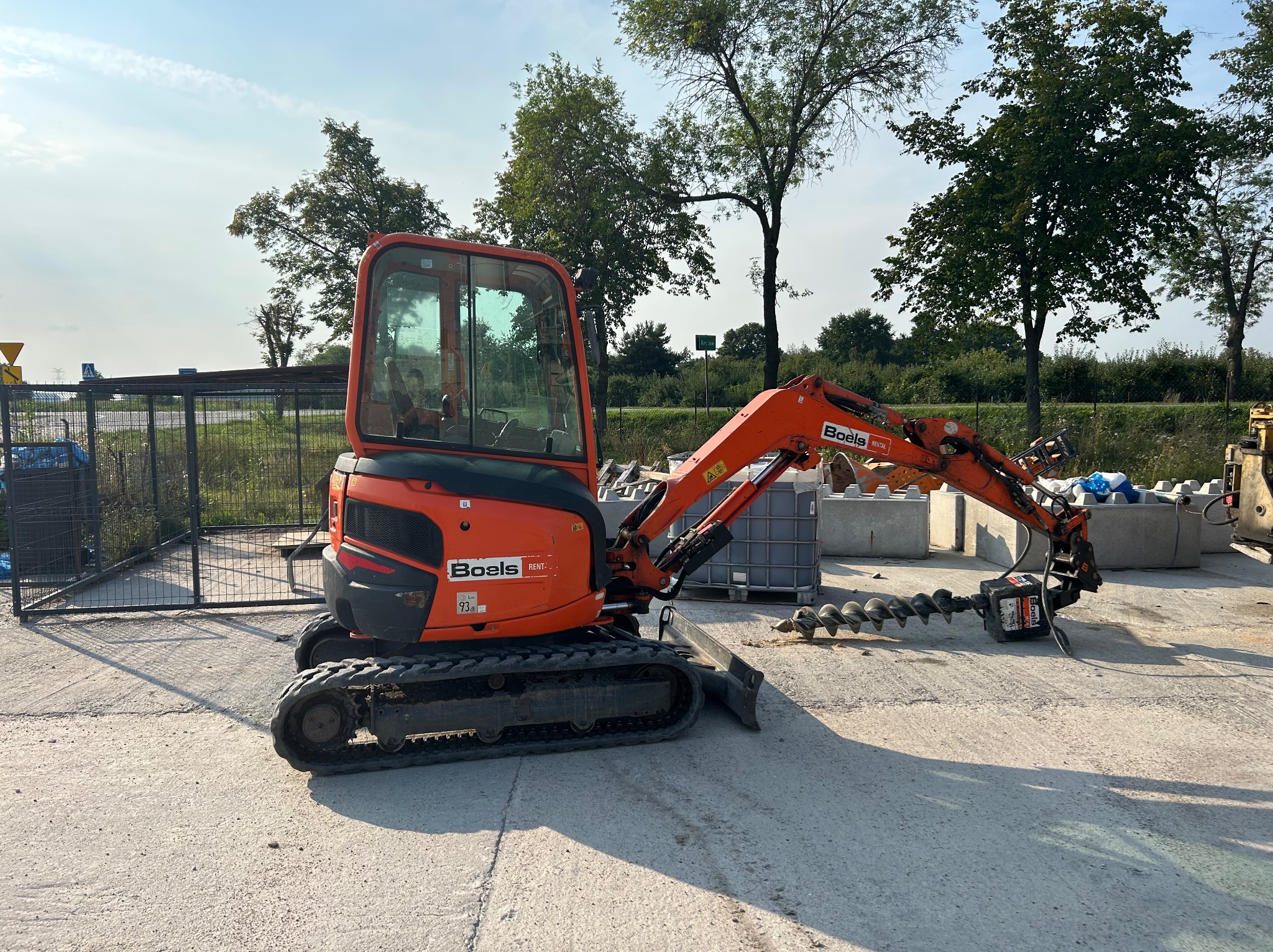 Pomarańczowa minikoparka gąsienicowa z wiertłem ślimakowym na betonowym podłożu, w tle drzewa i ogrodzenie, widoczne logo Boels Rental.