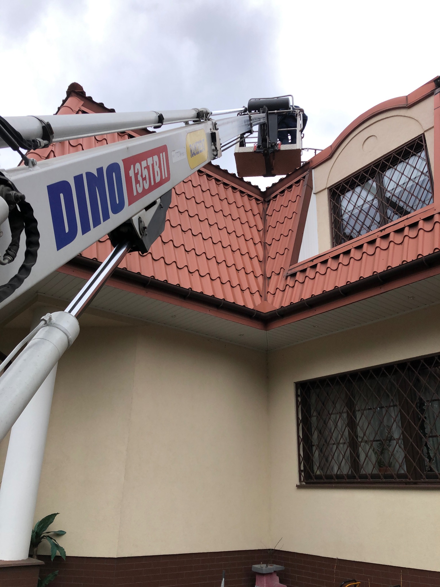 Praca na wysokościach przy użyciu podnośnika koszowego DINO 135TB II, widoczny fragment dachu z brązową dachówką i okno z kratą w domu o beżowej elewacji.