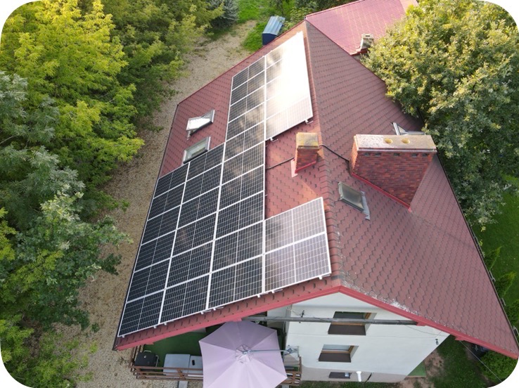 Widok z góry na dom z panelami fotowoltaicznymi na dachu z czerwonej dachówki bitumicznej, obok komin z cegły, otoczony zielenią drzew.