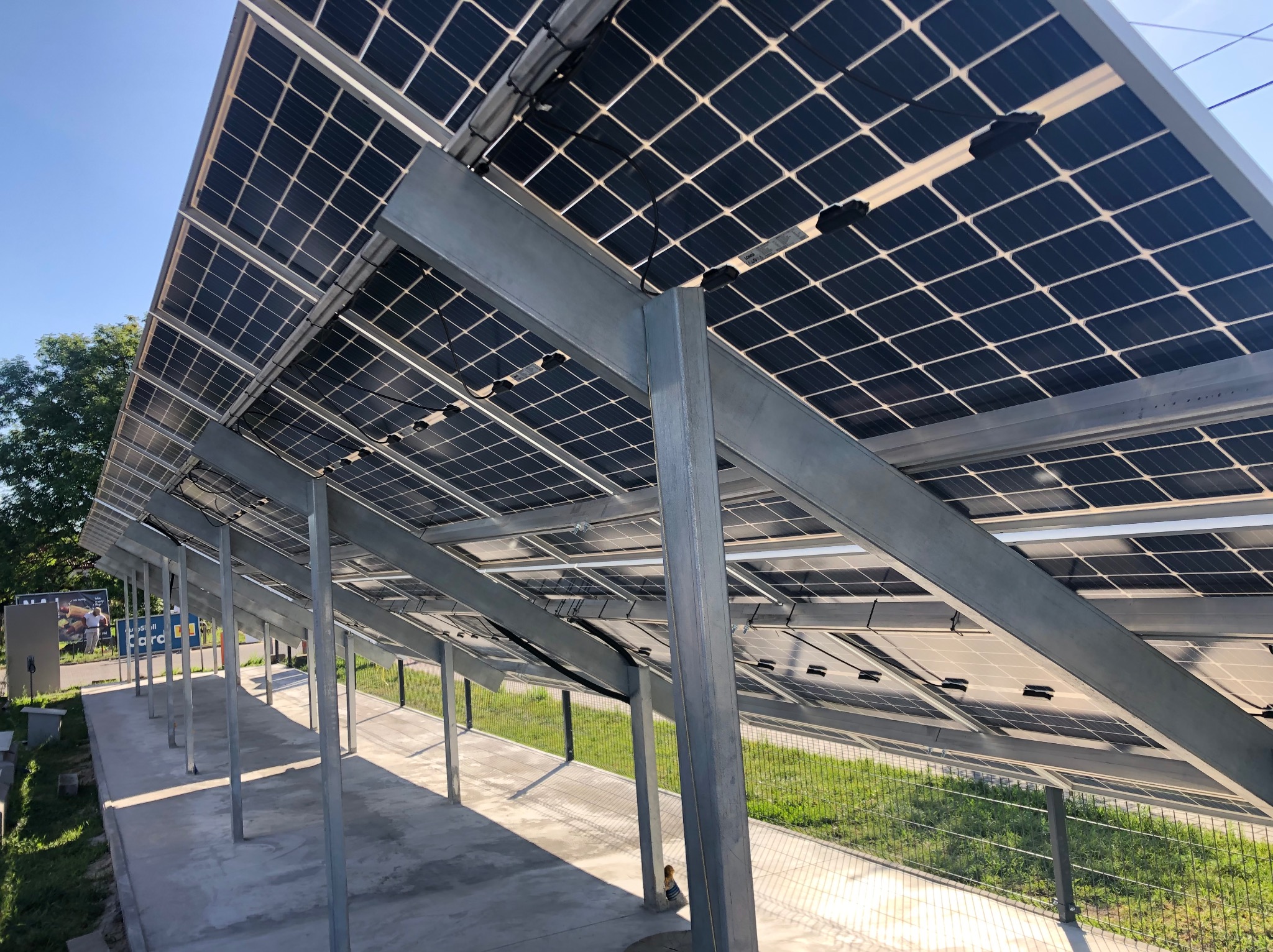 Widok spod spodu zadaszenia z paneli fotowoltaicznych, wspartych na metalowej konstrukcji, z widocznym fragmentem trawnika i betonowej nawierzchni.