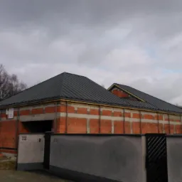 Dom w trakcie budowy z ciemnym, metalowym dachem na etapie wykończenia. Widoczne elementy konstrukcyjne i otoczenie budynku. Pochmurne niebo w tle.