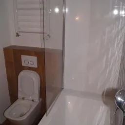 Nowoczesna łazienka z podwieszaną toaletą w zabudowie z drewnopodobnych paneli, szklanym parawanem nawannowym i grzejnikiem drabinkowym.