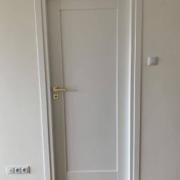 JAZZ BUD MICHAŁ BŁASZCZYŃSKI - Białe, nowoczesne drzwi wewnętrzne z prostą ramą, zamontowane w świeżo pomalowanym pomieszczeniu z drewnianą podłogą i gniazdkami elektrycznymi przy podłodze.