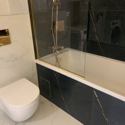 JAZZ BUD MICHAŁ BŁASZCZYŃSKI - Nowoczesna łazienka z białymi marmurowymi płytkami i czarną marmurową obudową wanny, złote akcenty na szklanym panelu prysznicowym i przycisku spłuczki, podwieszana toaleta.