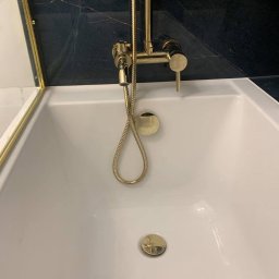 JAZZ BUD MICHAŁ BŁASZCZYŃSKI - Biała wanna z zamontowaną złotą armaturą i prysznicem na tle ciemnej ściany z marmurowym wzorem.