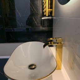 JAZZ BUD MICHAŁ BŁASZCZYŃSKI - Umywalka nablatowa w kształcie łzy z złotym rantem i baterią w kolorze złota na tle ściany wyłożonej czarnym marmurem ze złotymi żyłami oraz podświetlanego lustra.