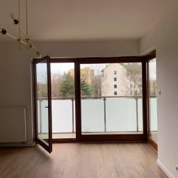 JAZZ BUD MICHAŁ BŁASZCZYŃSKI - Pusty pokój z drewnianą podłogą, otwartym balkonem z matową szybą, nowoczesną lampą sufitową i widokiem na budynek mieszkalny.