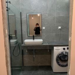 JAZZ BUD MICHAŁ BŁASZCZYŃSKI - Szara łazienka z kabiną prysznicową, umywalką nablatową, podświetlanym lustrem i pralką Samsung, widok zza ościeżnicy w trakcie prac wykończeniowych.