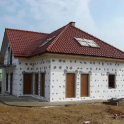 Dom jednorodzinny w trakcie ocieplania, z widoczną warstwą izolacyjną z charakterystycznymi plackami kleju. Czerwona dachówka i częściowo zamontowane okna.