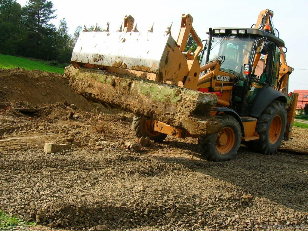 Żółta koparko-ładowarka CASE z łyżką pełną ziemi i darni na placu budowy, widok z boku na nierówny teren pokryty żwirem i ziemią.