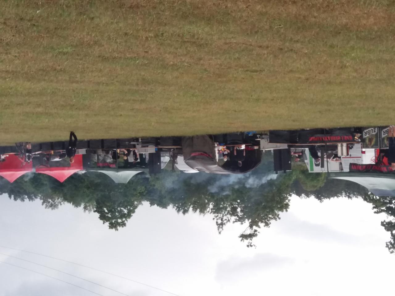 Catering na imprezie plenerowej z widocznymi stanowiskami grillowymi i namiotami, unoszący się dym z grilla, w tle drzewa i trawnik.