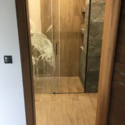 Nowoczesna kabina prysznicowa z czarnymi profilami i ozdobnym motywem na szkle, widoczna zza drewnianej ościeżnicy drzwi, z drewnopodobnymi płytkami na ścianach i podłodze.