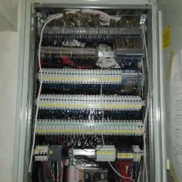 Otwarta skrzynka elektryczna z licznymi przewodami, bezpiecznikami i elementami sterującymi, widoczny miernik Voltcraft, schemat instalacji na ścianie.