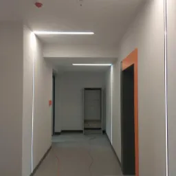 Korytarz biurowy z nowoczesnym oświetleniem LED: pionowe i poziome linie świetlne, pomalowane na biało ściany, szara podłoga, pomarańczowa futryna drzwiowa.