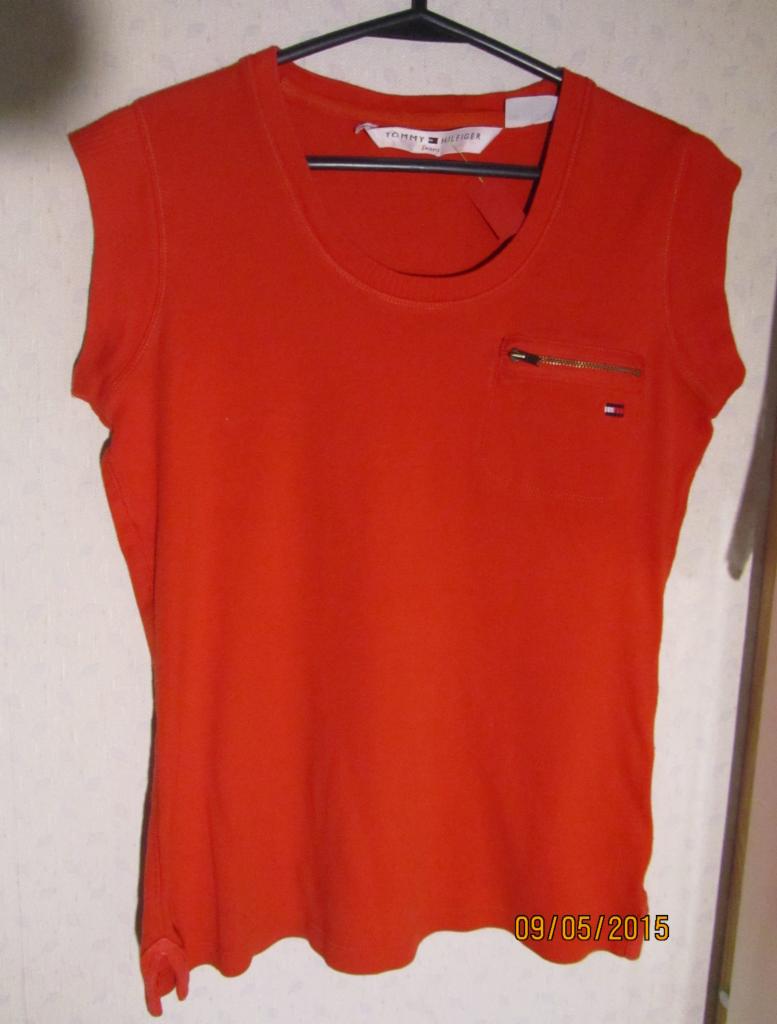 Pomarańczowa bluzka Tommy Hilfiger z krótkim rękawem, kieszonką na zamek i logo, wisząca na czarnym wieszaku. Zdjęcie z datą 09/05/2015.