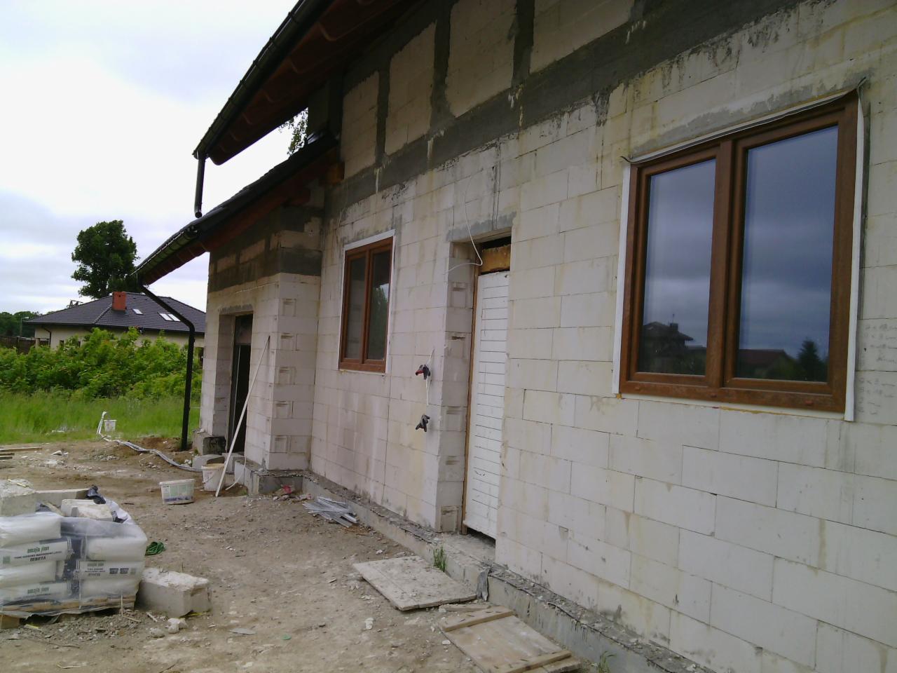 Surowy stan domu murowanego z oknami, workami cementu i widocznym zarysem dachu sąsiedniego budynku w tle. Widok z zewnątrz na budowę.