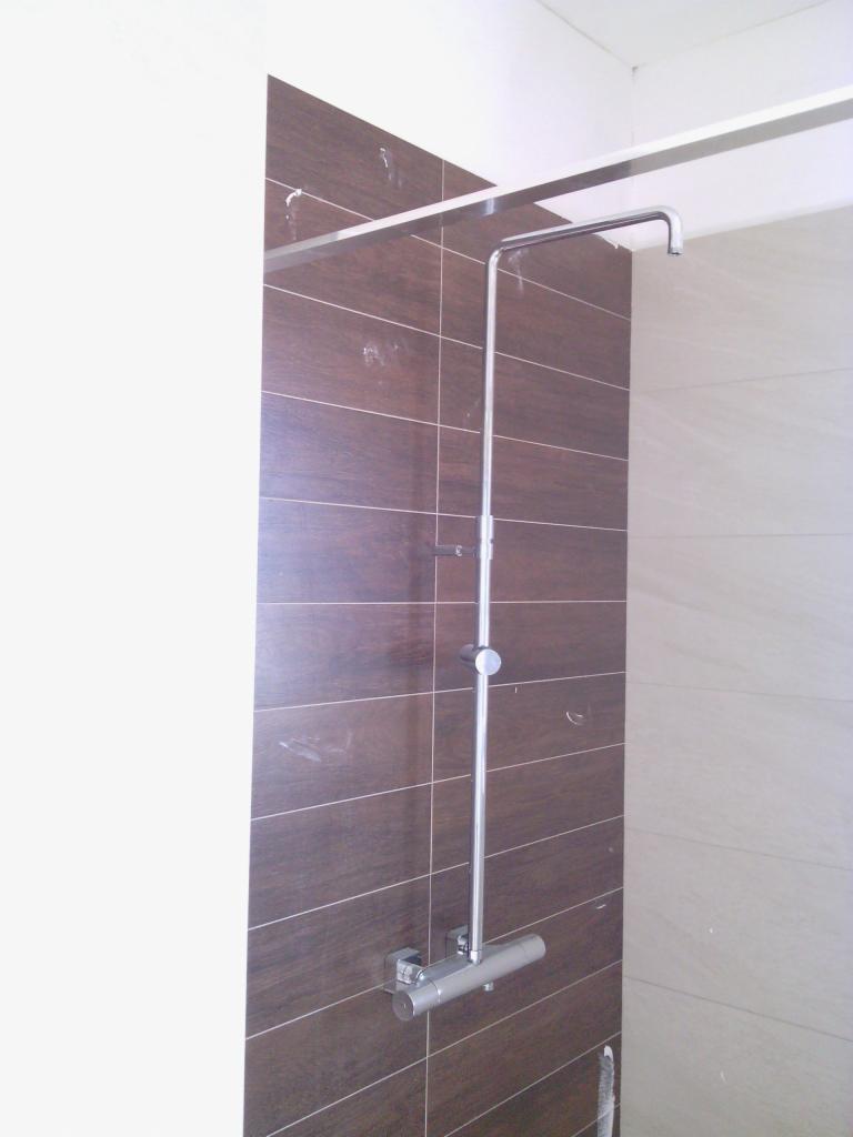 Nowoczesny prysznic z brązowymi płytkami w nowej łazience. Bateria termostatyczna i deszczownica zamontowana na ścianie. Wnętrze w trakcie wykończenia.