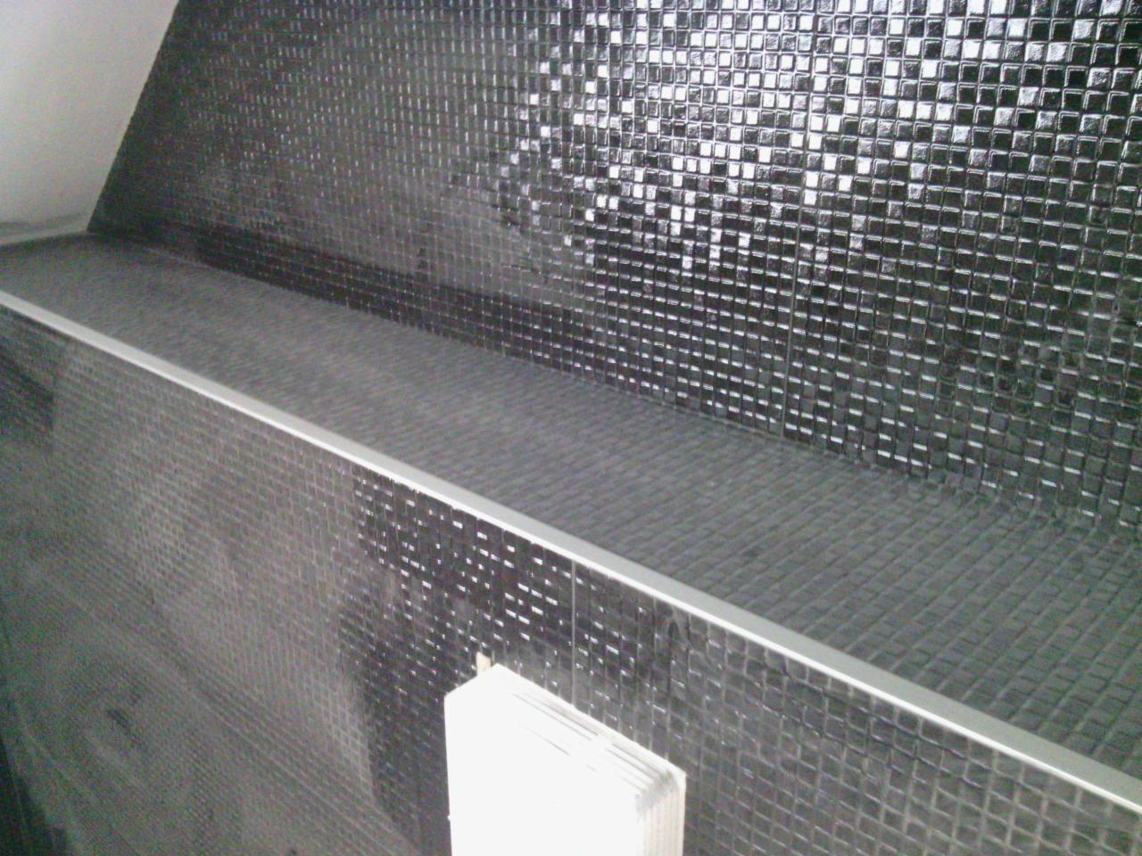 Ściana wyłożona błyszczącą, czarną mozaiką z drobnymi kwadratami, wykończona srebrną listwą. Widoczne fugi i refleksy światła.