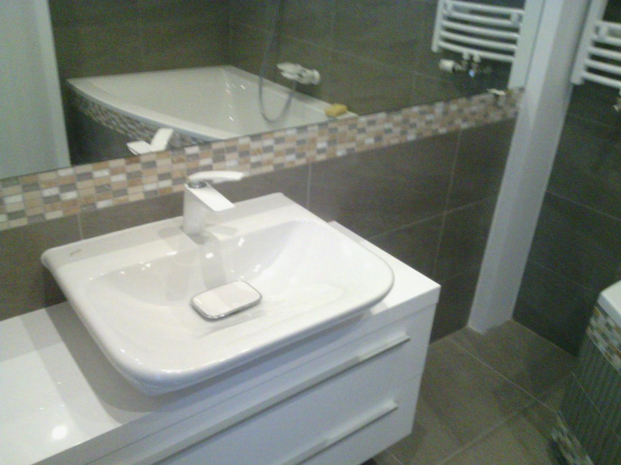 Nowoczesna łazienka z białą umywalką nablatową i baterią, z widocznym fragmentem wanny w lustrze. Szare płytki i białe meble łazienkowe.