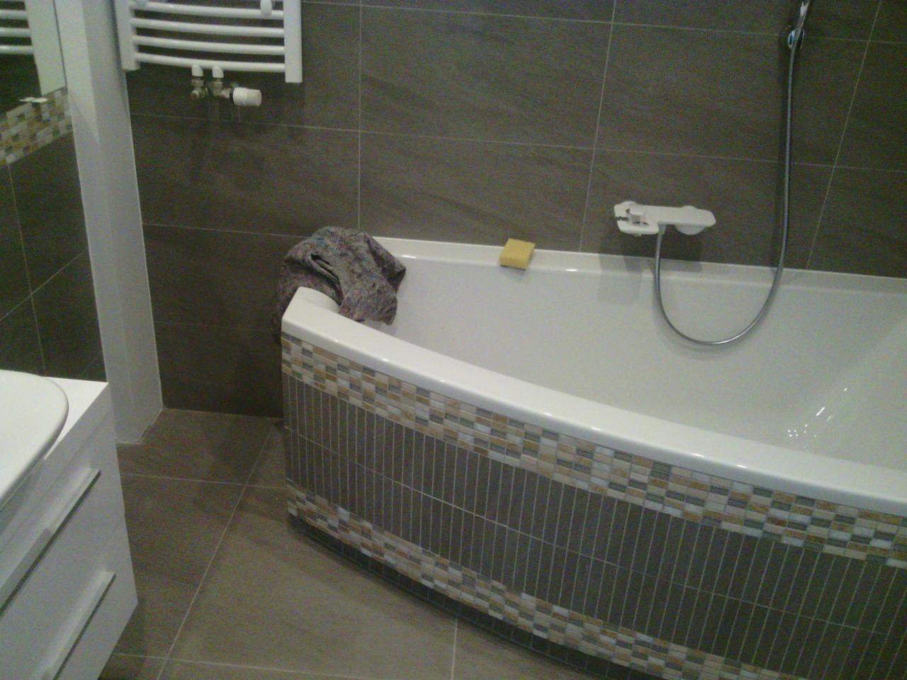 Wykończona łazienka: biała wanna z mozaiką, szare płytki ścienne i podłogowe, grzejnik drabinkowy. Widoczna gąbka i ręcznik na wannie.