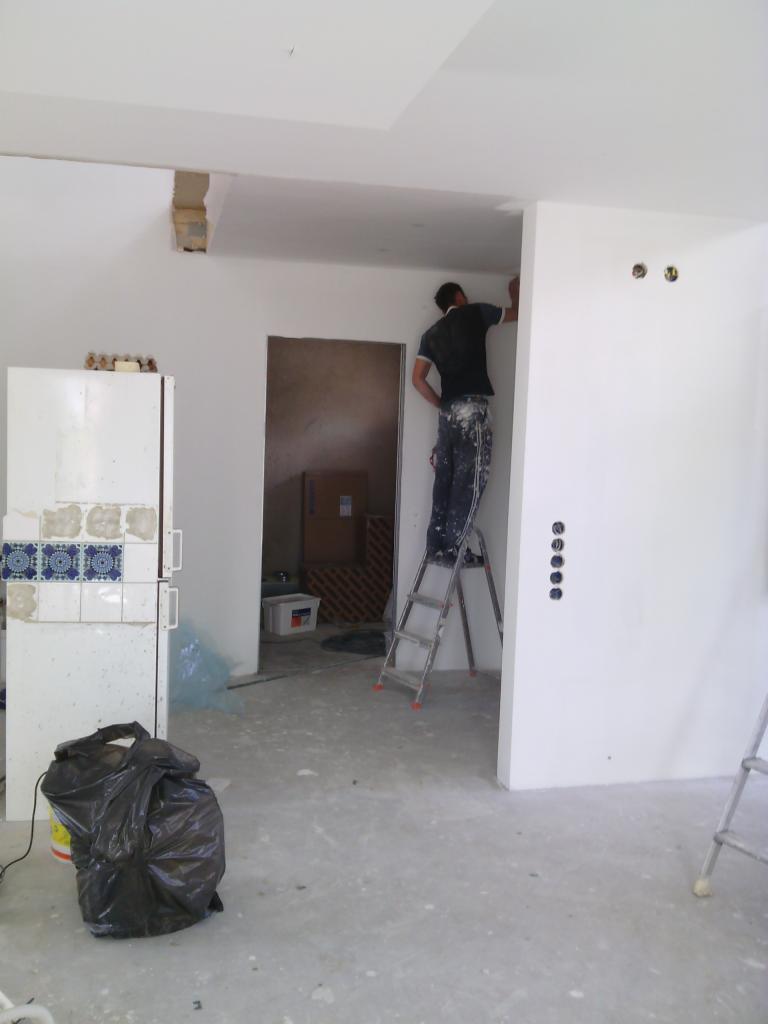 Pracownik maluje ścianę na biało, stojąc na drabinie w remontowanym wnętrzu. Widoczne przygotowania do prac wykończeniowych i instalacja elektryczna.