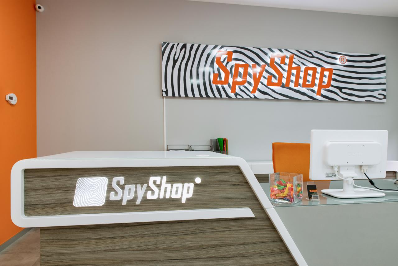Recepcja sklepu z logo SpyShop podświetlonym na drewnianym panelu, w tle baner ze wzorem zebry i napisem SpyShop, obok monitor i pojemnik ze słodyczami.