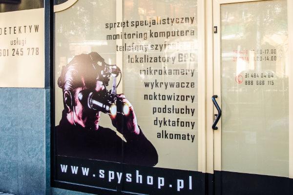 Witryna sklepu detektywistycznego z plakatem przedstawiającym mężczyznę z noktowizorem i listą oferowanego sprzętu: lokalizatory GPS, mikrokamery, wykrywacze, podsłuchy, dyktafony.