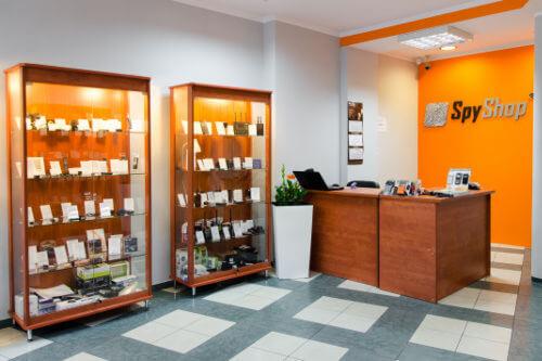 Wnętrze sklepu detektywistycznego 'Spy Shop' z dwoma witrynami wypełnionymi sprzętem szpiegowskim, biurkiem obsługi i pomarańczową ścianą z logo.