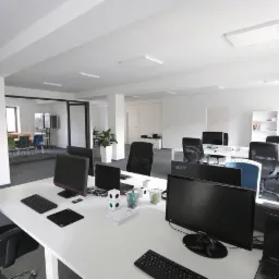 Jasne, przestronne biuro typu open space z białymi biurkami, komputerami i krzesłami biurowymi. W tle przeszklona sala konferencyjna i rośliny doniczkowe.