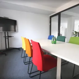 Nowoczesna sala konferencyjna z białym stołem, kolorowymi krzesłami i dużym ekranem LCD na mobilnym stojaku, widok na otwartą przestrzeń biurową przez szklane drzwi.