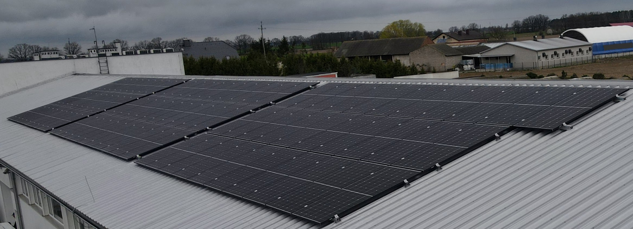 Panele fotowoltaiczne zamontowane na metalowym dachu budynku przemysłowego, widok z góry, pochmurne niebo w tle.