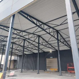 Wnętrze hali w trakcie budowy, widoczne stalowe słupy, konstrukcja dachu z płyt warstwowych, podnośnik koszowy oraz tablica informacyjna 'Brama 6'.