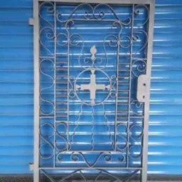 Metalowa, dekoracyjna brama z krzyżem w centrum, po obróbce, oparta o niebieską roletę.